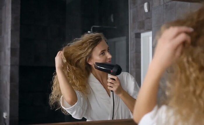 Woman blow-drying her curly hair in front of a mirror while preparing for a special event.