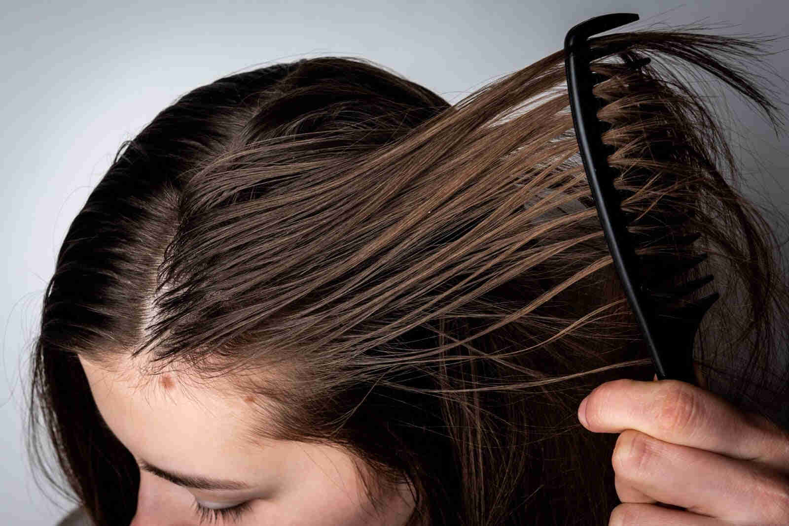 Close-up of a woman combing oily hair near the scalp with a black wide-tooth comb, showing visible greasiness and shine.