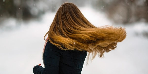 Woman with long, light brown hair walking outdoors in winter, her hair blowing in the cold air with snow in the background.