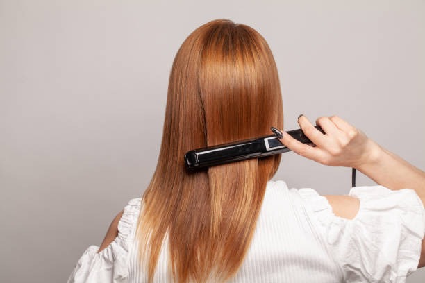 A woman using a flat iron hair straightener to smooth her long, shiny brown hair.